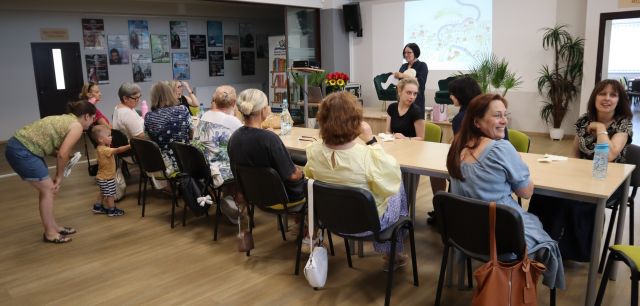 Na zdjęciu znajdują się kobiety siedzące na holu biblioteki przy złączonych stołach, przed nimi stoi kobieta z maskotką gęsi. W tle znajduje się prezentacja wyświetlana na ścianie, plakaty na ścianach.