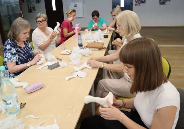 Na zdjęciu znajdują się kobiety przy złączonych stolikach i szyją swoje maskotki gęsi. Na stole znajdują się materiały, puch. W tle znajdują się plakaty na ścianach.