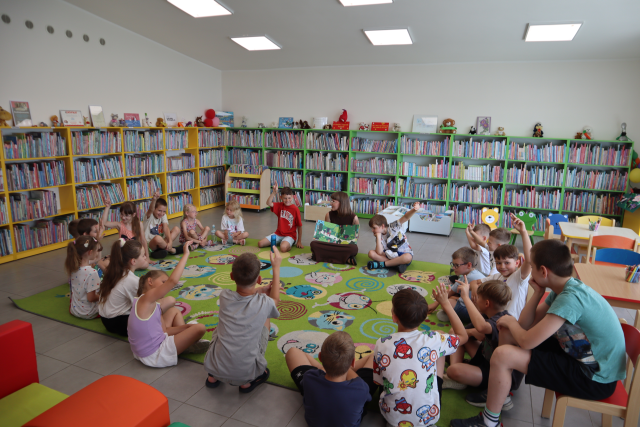 Na zdjęciu znajduje się około 20 dzieci na dywanie, część z nich ma podniesioną do góry rękę. Na środku siedzi kobieta z otwartą książką i pokazuje uczestnikom ilustracje. W tle znajdują się półki z książkami, po prawej stronie stoją dwa stoliczki z kredkami i krzesełka dookoła nich.