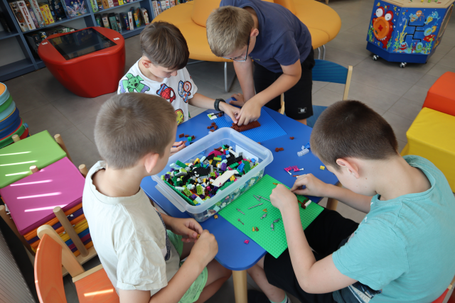 Na zdjęciu znajduje się czwórka chłopców przy stoliku i wspólnie układają klocki LEGO na dwóch podkładkach, na stoliku leży również plastikowy pojemnik z klockami. W tle znajdują się małe pufy, stolik interaktywny, okrągła kanapa oraz półki z grami.