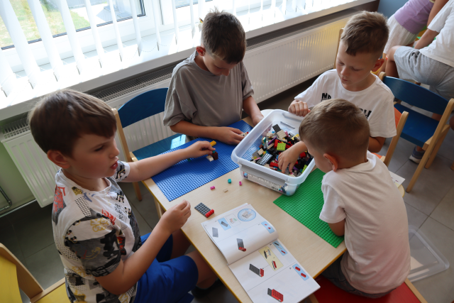 Na zdjęciu znajduje się czwórka chłopców, którzy budują z klocków LEGO na zielonej i niebieskiej podkładce. Klocki znajdują się w plastikowym pojemniku i jeden z chłopców szuka w nim klocków. W tle znajduje się grzejnik, okna oraz po prawej stronie dwójka dzieci przy sąsiednim stoliku.