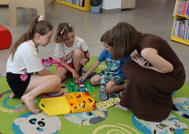Na zdjęciu znajdują się dwie dziewczynki oraz jeden chłopiec i wspólnie układają klocki LEGO na dywanie, obok nich kuca kobieta i przygląda się ich pracy. Na środku znajduje się również walizeczka z klockami LEGO. W tle znajdują się półki z książkami i nogi od stolika.