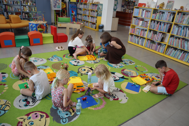 Na zdjęciu znajduje się ośmioro dzieci w parach na dywanie i układają klocki LEGO, które znajdują się w plastikowych walizeczkach. Przy jednej grupie kuca kobieta i przygląda się ich pracy. W tle znajdują się półki z książkami, pufy ułożone w pociąg, okrągła kanapa oraz stolik i miejsce do zabawy.