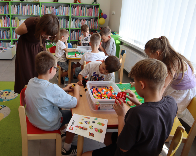 Na zdjęciu w centrum znajduje się czwórka dzieci przy stoliku i układają klocki LEGO, chłopiec po lewo pokazuje kobiecie nachylającej się obok niego swoją pracę. Za nimi w tle znajduje się stolik z czwórką chłopców, którzy również układają klocki LEGO, półki z książkami i zasłonięte okna po prawej stronie.