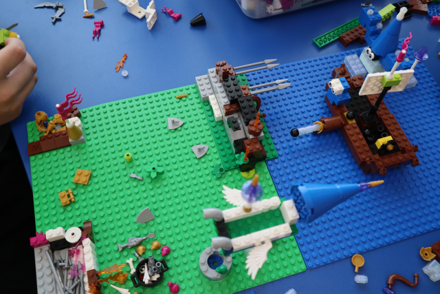 Na zdjęciu znajduje się konstrukcja z LEGO na dwóch podkładkach, zielonej i niebieskiej przedstawia statek, rakietę, barierę między jedną a drugą planszą. Praca leży na niebieskim stoliku a dookoła leżą klocki.
