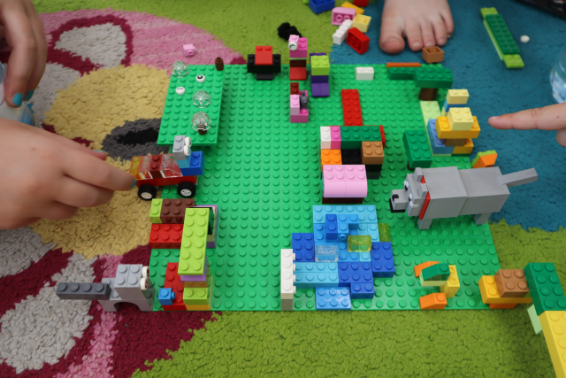 Na zdjęciu znajduje się makieta zbudowana z klocków LEGO przedstawiająca wodę, samochód, park oraz wilka, dookoła pracy, która znajduje się na dywanie leżą klocki. Po lewej stronie znajduje się ręka dziewczynki, która poprawia auto, a po prawej palec dziecka, które wskazuje na element makiety.
