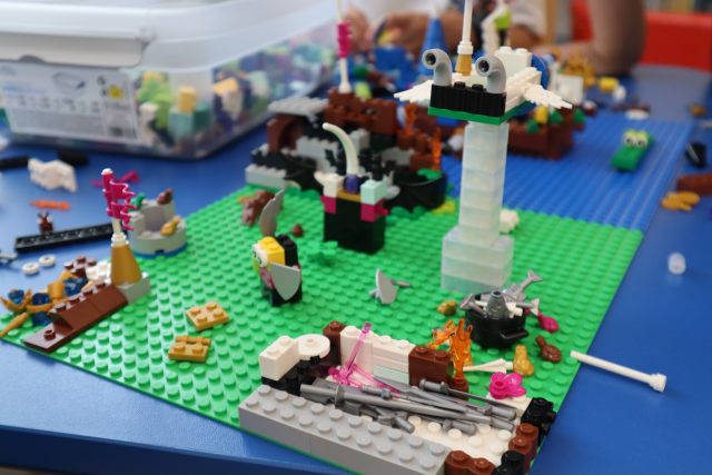 Na zdjęciu znajduje się makieta z klocków LEGO na zielonej płytce przedstawiająca kolumnę, ognisko. Dookoła leżą klocki LEGO na niebieskim stoliku oraz plastikowe pudełko z klockami LEGO.