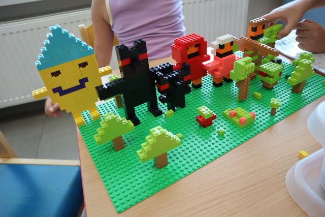 Na zdjęciu znajduje się makieta z klocków LEGO, na dwóch zielonych podkładkach, znajdują się na niej drzewam dwa wilki jeden mniejszy drugi większy, czerwony kapturek, babcia, leśniczy oraz uśmiechnięte słońce z nagimi, rękoma oraz niebieską czapką. Praca leży na drewnianym stoliku, za którym siedzi dziewczynka, a po prawej stronie dziecko, którego widoczna jest ręka poprawia jedno z drzew.