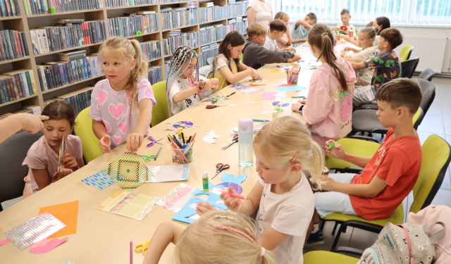 Na zdjęciu znajdują się dzieci przy złączonych stolikach na których leżą kolorowe kartki, cekiny, kredki, nożyczki. Na końcu stołu przy uczestnikach stoi kobieta. W tle znajdują się półki z książkami oraz okna.