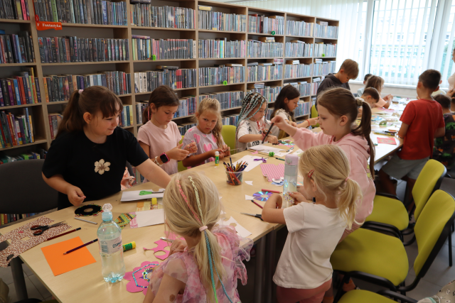 Na zdjęciu znajdują się dzieci przy złączonych stolikach i za pomocą kolorowych kartek, cekinów ozdabiają płyty. W tle znajdują się półki z książkami i okna.