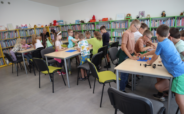 Na zdjęciu znajdują się trzy rzędy złączonych stolików przy których siedzą dzieci i w grupach układają z klocków LEGO. W tle znajdują się półki z książkami.