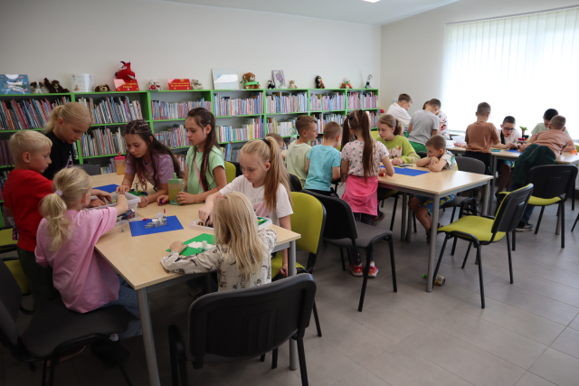 Na zdjęciu znajdują się dzieci i układają klocki LEGO przy złączonych stolikach w trzech rzędach. W tle znajdują się półki z książkami i okna po prawej stronie.