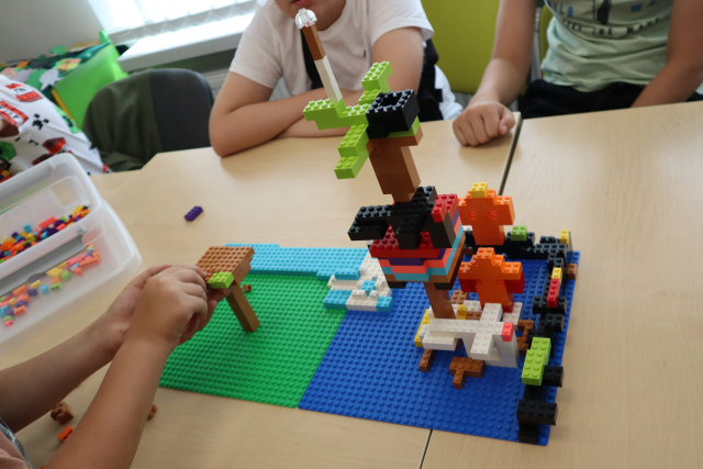 Na zdjęciu znajduje się konstrukcja z LEGO przedstawiająca domek na drzewie dookoła jest woda, brama obronna. Przy stoliku siedzą chłopcy, po lewej stronie jeden z uczestników, którego widoczne są tylko ręce doczepia klocek do drzewa, obok niego znajduje się plastikowy pojemnik z klockami. Praca leży na złączonych stolikach.