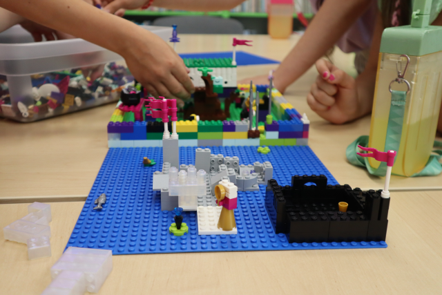 Na zdjęciu znajduje się zbudowany z klocków LEGO zamek na niebieskiej podkładce na złączonych stołach. W tle znajdują się ręce uczestniczek zajęć które dokładają klocki do swojej pracy, która przedstawia las, obok nich leży pudełko z klockami oraz po prawej stronie butelka z piciem.