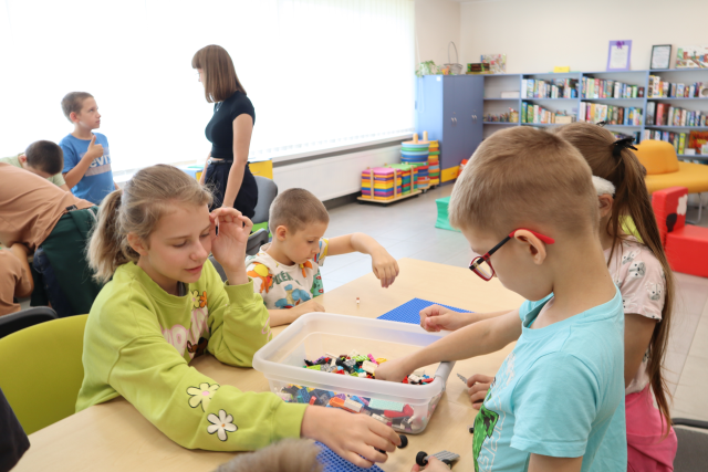 Na zdjęciu w centrum znajduje się czwórka dzieci, którzy wspólnie budują klocków LEGO znajdujących się na złączonych stolach. W tle znajduje się kobieta stojąca z chłopcem obok stolika obok, przy którym chłopcy budują z klocków. W tle znajdują się również okna, półki z grami, pufy.
