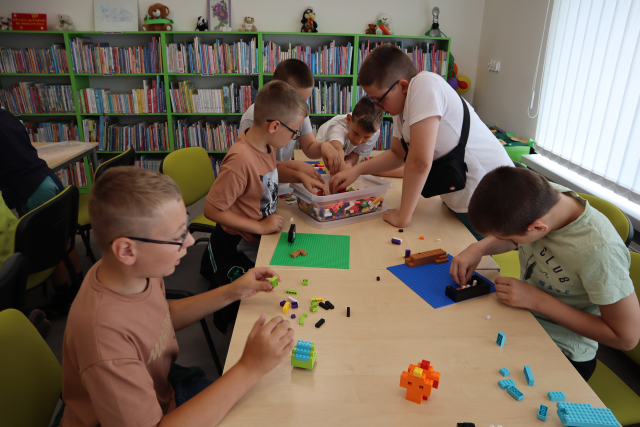 Na zdjęciu znajduje się szóstka chłopców, którzy budują z klocków LEGO przy złączonych stołach. W tle znajdują się półki z książkami.