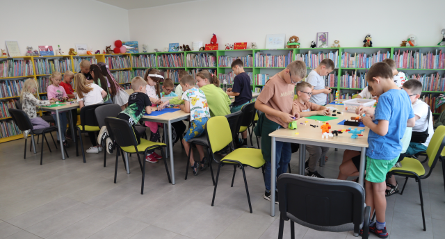 Na zdjęciu znajduje się około 20 dzieci przy złączonych stolikach w trzech rzędach i wspólnie układają z klocków LEGO. W tle znajdują się półki z książkami.