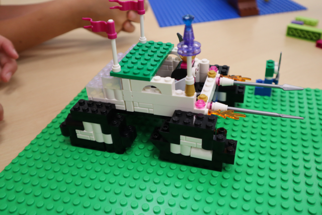 Na zdjęciu znajduje się zbudowany z klocków LEGO pojazd na zielonej podkładce na stole, posiada dwie flagi, kierowcę zbudowanego z klocków, na przodzie samochód posiada wystawione włócznie oraz płomienie ognia.