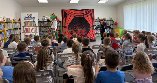 Na zdjęciu znajdują się dzieci siedzące na widowni w sali bibliotecznej, przed nimi znajduje się przebrana kobieta z igłą w ręce oraz sznurkiem i ciągnie za jego pomocą mężczyznę przebranego za smoka. W tle znajduje się tablica z czerwoną kurtyną, baner z logiem biblioteki i książkami oraz półki z książkami.
