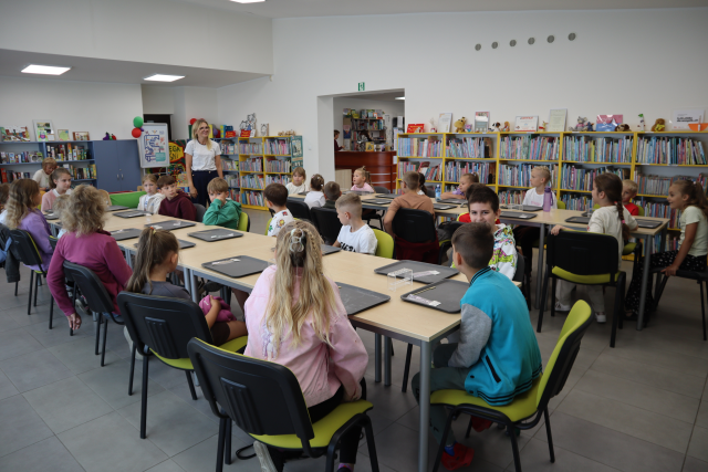 Na zdjęciu znajduje się dwadzieścia sześć dzieci przy złączonych stolikach w dwóch rzędach, przed nimi na stolikach leżą szare tacki. Pomiędzy stołami na początku stoi kobieta. W tle znajdują się półki z książkami.