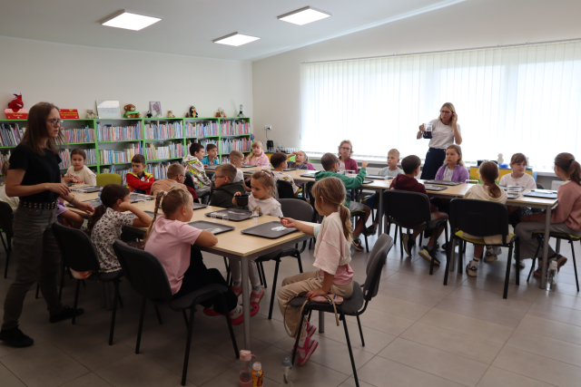 Na zdjęciu znajdują się dzieci przy złączonych stołach w dwóch rzędach, przed sobą mają szare tacki, po lewej stronie przy stoliku stoi kobieta, a kobieta stojąca po prawo trzyma dzbanek z herbatą. W tle znajdują się półki z książkami i okna.