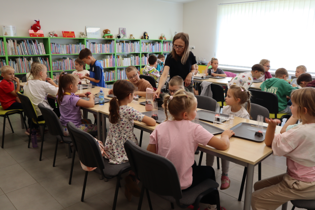Na zdjęciu znajdują się dwa rzędy złączonych stołów przy których siedzą dzieci i robią własną kompozycje herbat, pomiędzy rzędami stoi kobieta i podaje chłopcu jeden ze składników. W tle znajdują się półki z książkami oraz okna.