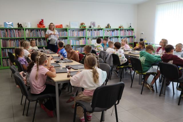 Na zdjęciu znajdują się dzieci przy złączonych w dwóch rzędach stolikach, przed sobą mają szare podkładki, Pomiędzy stolikami na końcu stoi kobieta z workiem herbaty. W tle znajdują się półki z książkami.