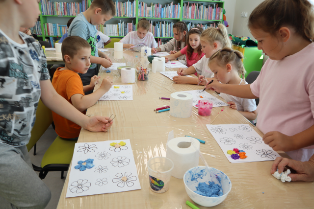 Na zdjęciu znajdują się dzieci przy złączonych stołach, które są owinięte folią. Uczestnicy zajęć ozdabiają wydrukowane kwiaty za pomocą masy z kleju, farb i papieru toaletowego, który znajduje się na stole. W tle znajdują się półki z książkami.