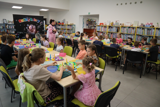 Na zdjęciu znajdują się dzieci przy złączonych stolikach w dwóch rzędach, przed nimi znajdują się przybory potrzebne do zajęć. Pomiędzy stolikami stoi kobieta, a w tle znajdują się półki z książkami i płachta z napisem Bańki i Kolory.