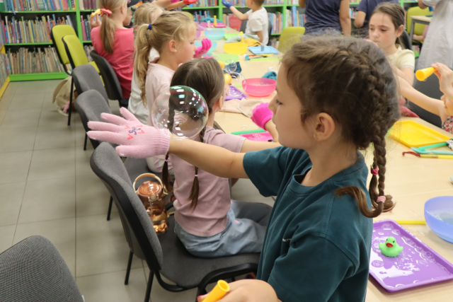Na zdjęciu z centrum znajduje się dziewczynka stojąca bokiem, ma wystawioną rękę, na której ma rękawiczkę i bańkę mydlaną. W tle znajdują się złączone stoliki z uczestnikami zajęć oraz półki z książkami.