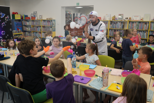 Na zdjęciu znajdują się dwa rzędy złączonych stolików, przy których siedzą dzieci i łapią na dłonie z rękawiczką bańki puszczane przez mężczyznę. W tle znajdują się półki z książkami,