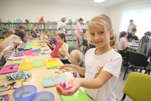 Na zdjęciu w centrum znajduje się dziewczynka z wystawioną ręką w której trzyma bańkę z małą zabawką. W tle znajdują się dzieci przy złączonych stolikach, mężczyzna i dwie kobiety oraz półki z książkami.