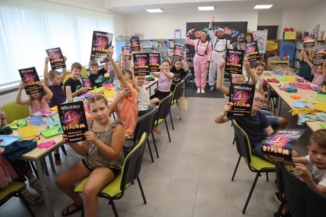 Na zdjęciu znajdują się dzieci przy złączonych w dwóch rzędach stołach i podnoszą do góry rękę z dyplomem z Baniek i Kolorów. Na stolikach leża miseczki, tacki i ręczniki z pomiędzy stołami stoją dwie kobiety i jeden mężczyzna i machają do zdjęcia. W tle znajdują się okna i półki z książkami.