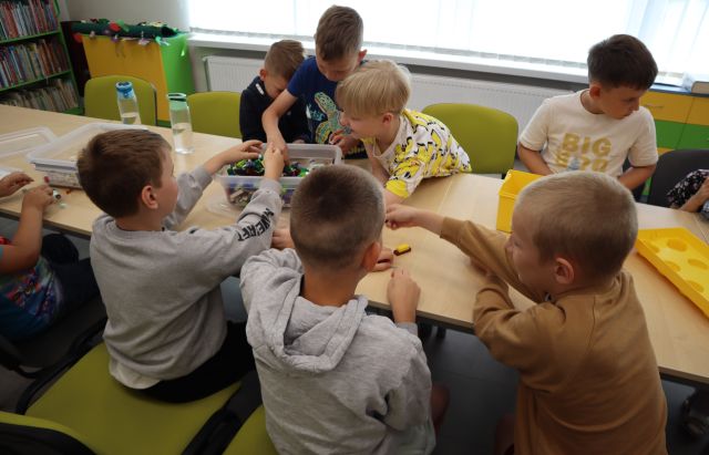 Na zdjęciu znajduje się osiem chłopców po obu stronach złączonych stołów przy których układają klocki LEGO oraz szukają elementów w pudełkach. W tle znajdują się okna.