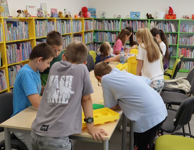 Na zdjęciu znajdują się dzieci przy złączonych stołach i układają klocki LEGO, które znajdują się w pudełkach. W tle znajdują się półki z książkami.