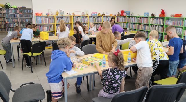 Na zdjęciu znajdują się dwa rzędy złączonych stołów, przy których stoją dzieci i układają klocki LEGO. W tle znajdują się półki z książkami.