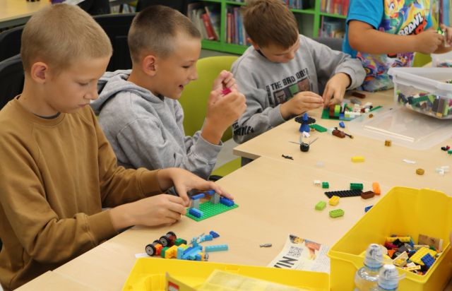 Na zdjęciu znajduje się czterech chłopców, trójka stoi przy złączonych stołach, a jeden stoi i układają klocki LEGO, przed nimi leżą klocki oraz pudełka. W tle znajdują się półki z książkami.