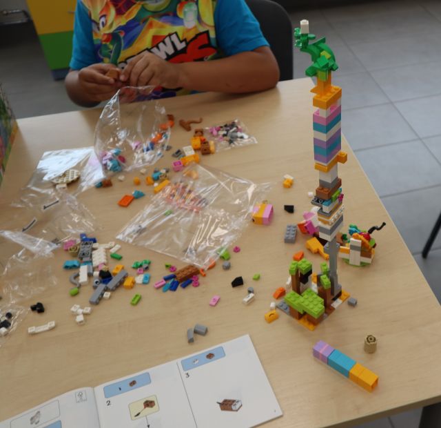 Na zdjęciu znajduje się stolik, na którym leżą klocki LEGO oraz jej zbudowana wieża, przy stoliku siedzi chłopiec i składa klocki.