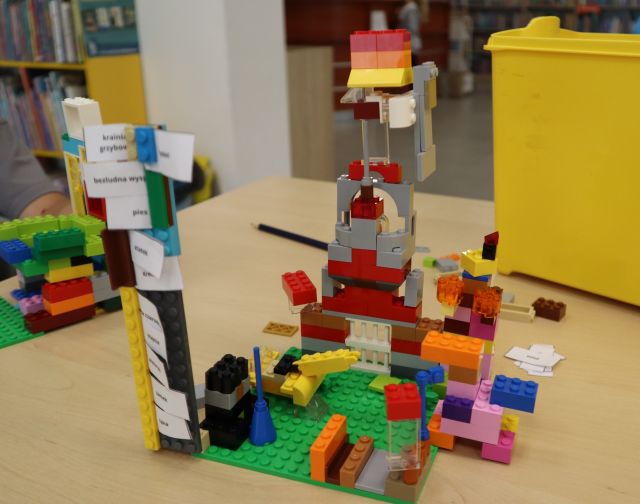 Na zdjęciu znajduje się stojąca na stoliku konstrukcja z klocków LEGO przedstawiająca wieże oraz znak, gdzie włożone są papierowe karteczki z napisami. W tle znajdują się półki z książkami.
