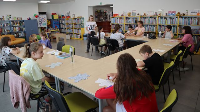 Na zdjęciu znajdują się dzieci przy złączonych stolikach w podkowę, przed sobą mają nożyczki i materiały w kształcie misia. Przed nimi stoi kobieta. W tle znajdują się półki z książkami.