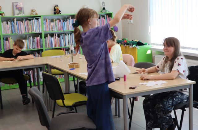 Na zdjęciu w centrum znajdują się dwie dziewczynki, jedna z nich stoi i nawleka nitkę, obok nich przy złączonych stołach siedzi dziewczynka oraz chłopiec i szyją misia. W tle znajdują się półki z książkami.