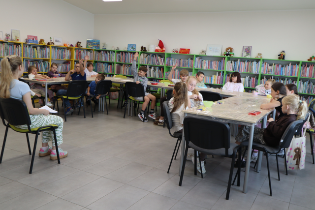 Na zdjęciu znajdują się dzieci przy złączonych w podkowę stołach, na środku siedzi kobieta i czyta im książkę. Część dzieci podnosi do góry rękę. W tle znajdują się półki z książkami.