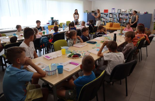 Na zdjęciu znajdują się dzieci przy złączonych w dwóch rzędach stolikach, na których leżą tacki, miseczki, talerze z owocami. Przed stołami stoją trzy kobiety. W tle znajdują się półki z książkami i okna.