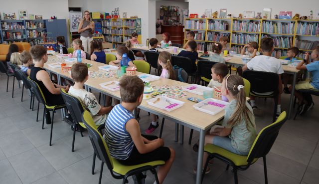 Na zdjęciu znajdują się dzieci przy złączonych w dwóch rzędach stołach, przed sobą mają tacki, talerze z owocami. Na środku między stolikami stoi kobieta. W tle znajdują się półki z ksiażkami.