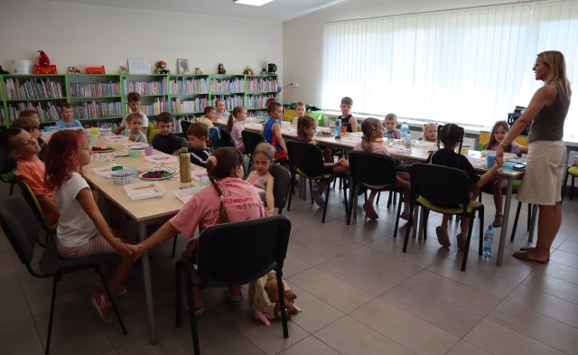 Na zdjęciu znajdują się dzieci przy złączonych w dwóch rzędach stolikach i trzymają się za ręce, przy drugim stoliku stoi kobieta i trzyma się razem z dziećmi za ręce. W tle znajdują się półki z książkami i okna.