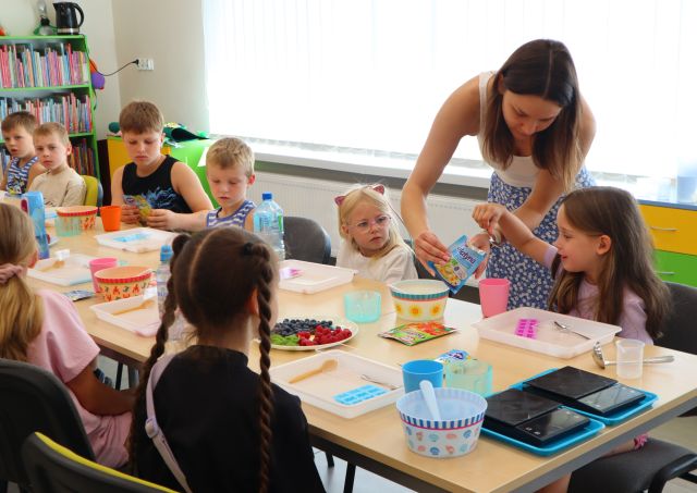Na zdjęciu znajdują się dzieci przy złączonych stolikach, przed nimi leżą tacki, miseczki, talerz z owocami. Przy jednej dziewczynce nachyla się kobieta z opakowaniem, a dziewczynka nasypuje go do kubeczka. W tle znajdują się okna oraz półki z książami.
