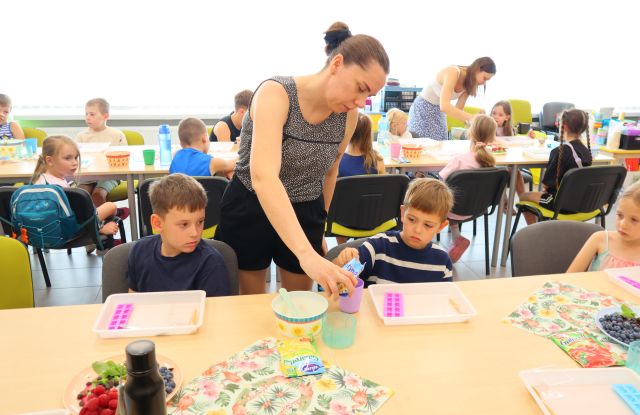 Na zdjęciu w centrum znajduje się dwóch chłopców przy złączonych stolikach, jeden z nich nasypuje żelatynę z trzymanej przez kobietę obok saszetki. Na stoliku stoją tacki, miseczki owoce. Po prawej stronie przy stoliku siedzi również dziewczynka. W tle znajduje się drugi rząd stolików przy których siedzą dzieci oraz nachyla się kobieta.