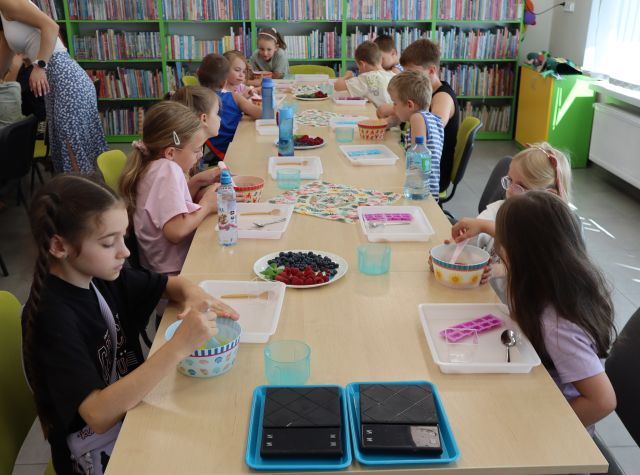 Na zdjęciu znajdują się dzieci przy złączonym stoliku i mieszą galaretkę w miseczkach. Na stolikach znajdują się tacki, łyżki, owoce, bidony i dwie wagi na początku stołu. Po lewej stronie znajduje się kobieta. W tle znajdują się półki z książami.