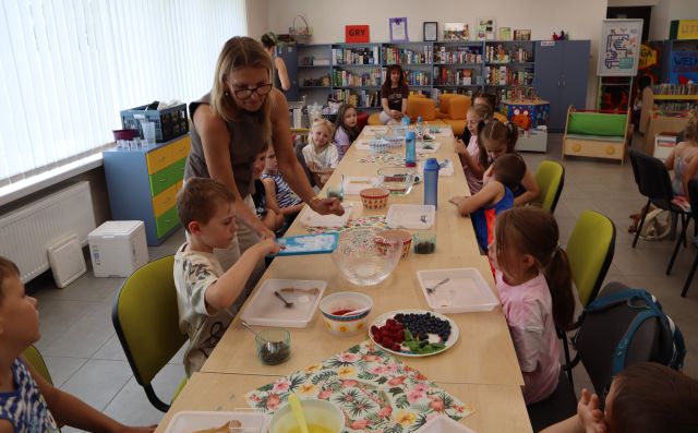 Na zdjęciu znajdują się złączone stoliki przy których siedzą dzieci, na stolikach leżą tacki, miski, miseczki, owoce. Po lewej stronie stoi kobieta podaje chłopcu na tacce suchy lód do dotknięcia. W tle znajdują się półki z książkami, po lewej stronie okna.