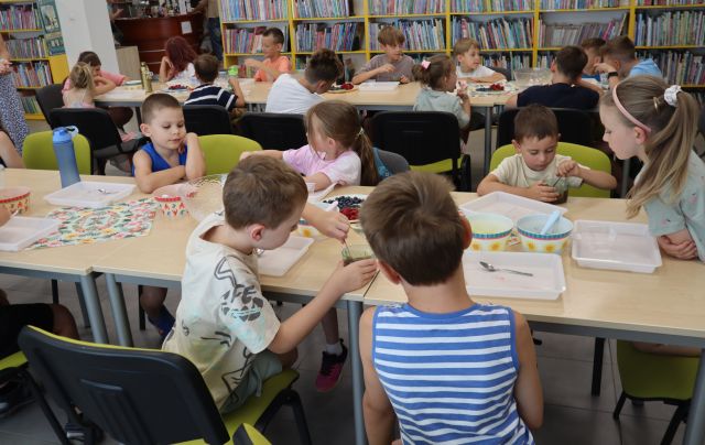 Na zdjęciu znajdują się dzieci przy złączonych stolikach w dwóch rzędach i mieszają czekoladę w pojemniczkach. Na stolikach znajdują się tacki, miseczki, owoce, serwetki. W tle znajdują się półki z książami.